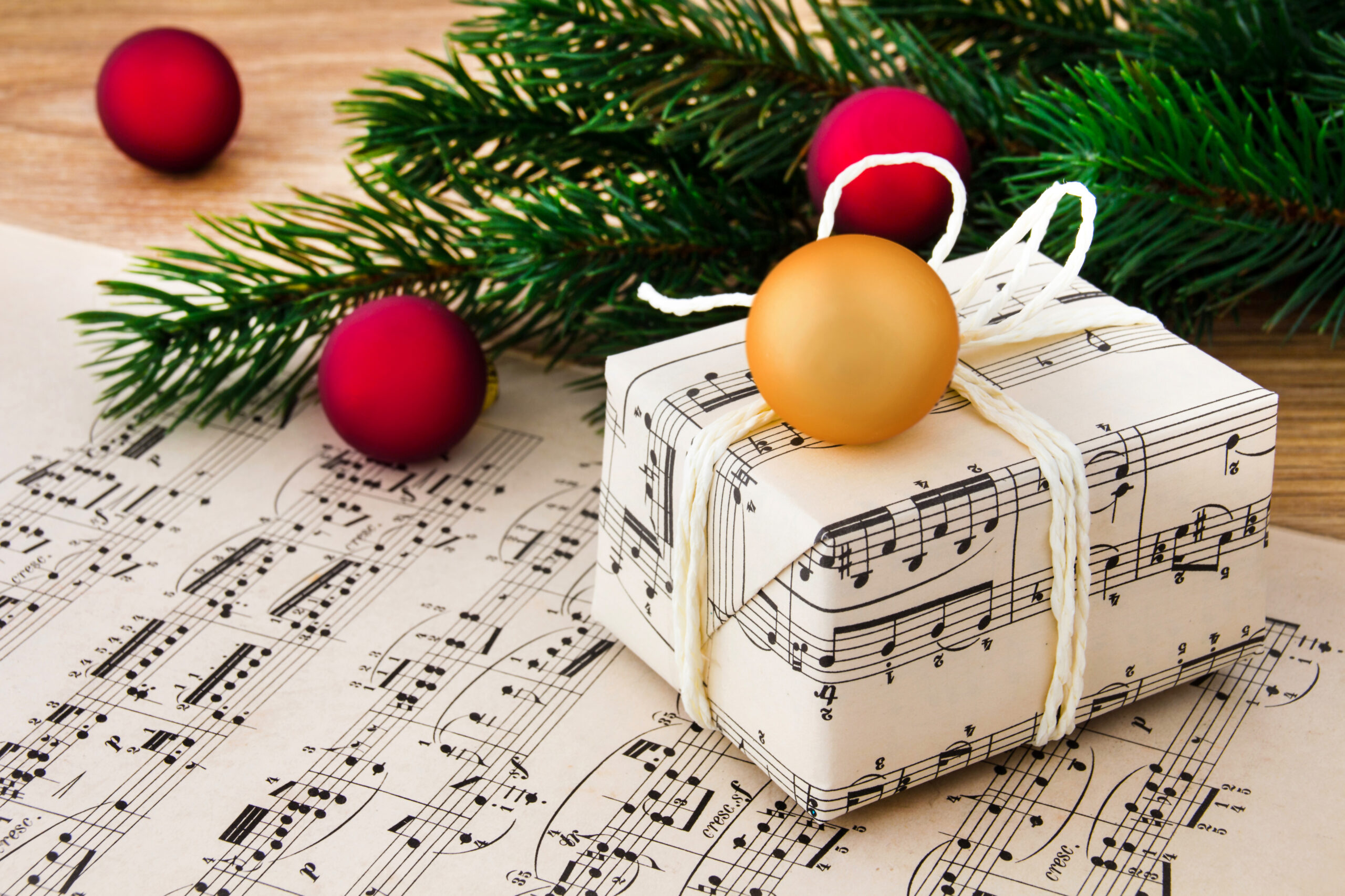 Ein eingepacktes Weihnachtspaket mit weißer Schnur auf einem Notenblatt, darauf liegt eine goldene Weihnachtskugel. Im Hintergrund liegt ein Tannenzweig mit 3 roten Kugeln